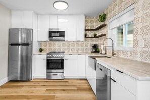 A modern kitchen with white cabinetry, stainless steel appliances, and patterned wallpaper. - Beachside Bungalow - Unit A | 2BR, 8-Min Walk to Beach, Patio & BBQ Grills 🌴 (Cape Canaveral)