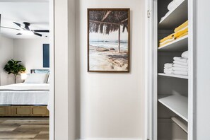 A bedroom with a bed, a ceiling fan, a wall-mounted painting of a beach scene, and a storage area with towels. - Beachside Bungalow - Unit A | 2BR, 8-Min Walk to Beach, Patio & BBQ Grills 🌴 (Cape Canaveral)