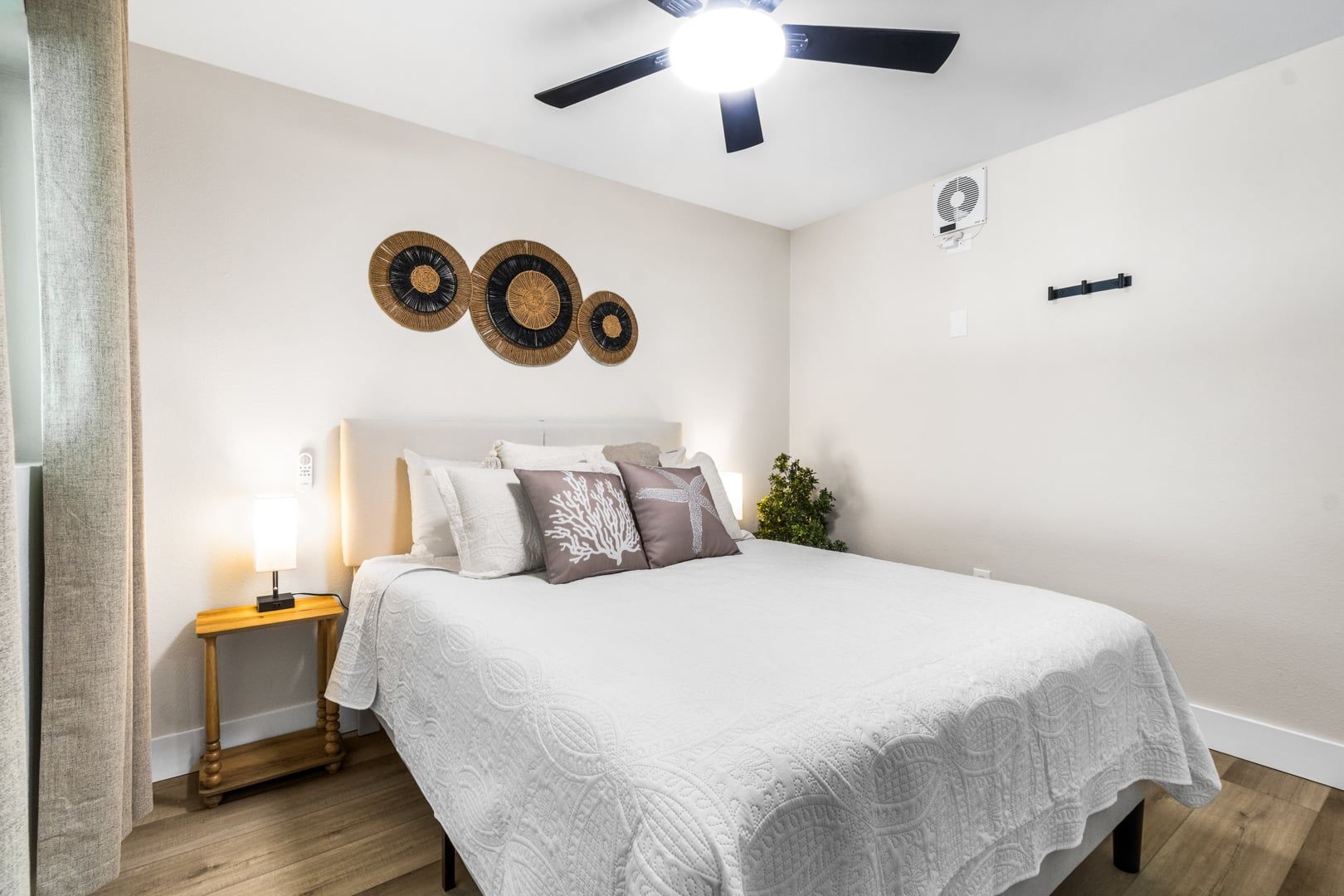 A bedroom with a bed, bedside table, lamp, wall decor, ceiling fan, and a small plant.