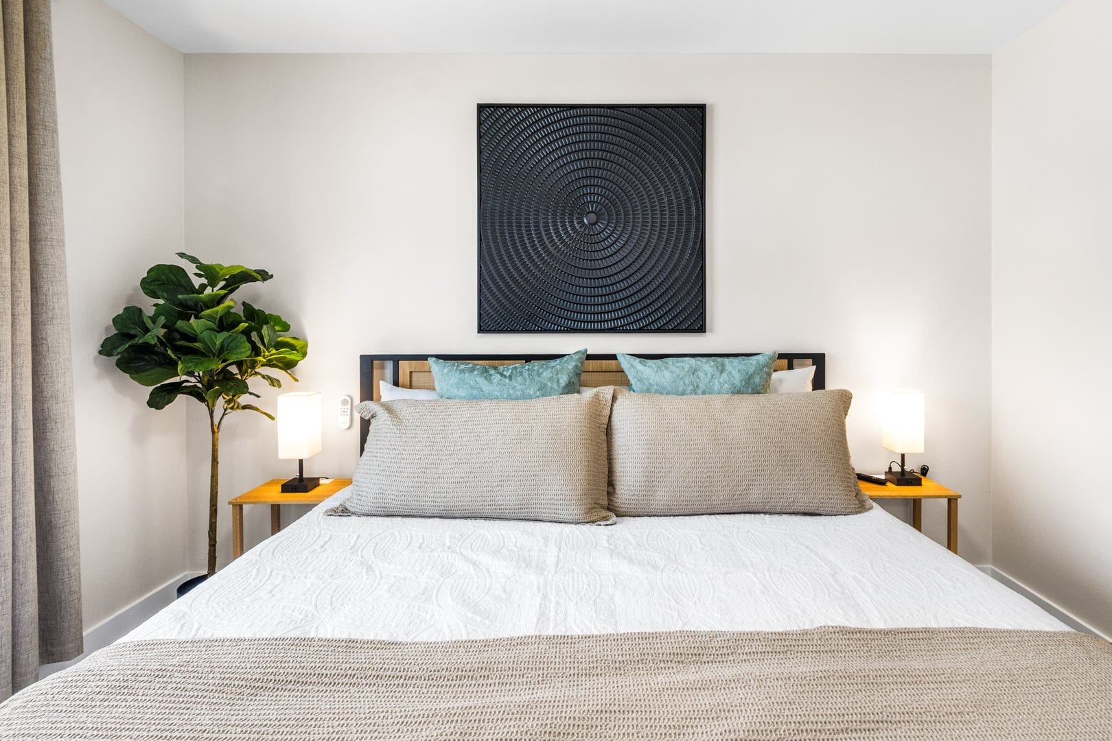 A neatly made bed with a white comforter and two pillows, flanked by bedside tables with lamps, a wall-mounted artwork, and a potted plant.