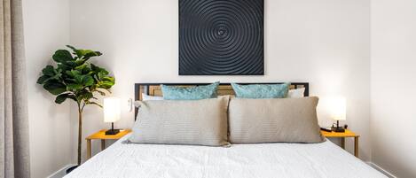 A neatly made bed with a white comforter and two pillows, flanked by bedside tables with lamps, a wall-mounted artwork, and a potted plant.
