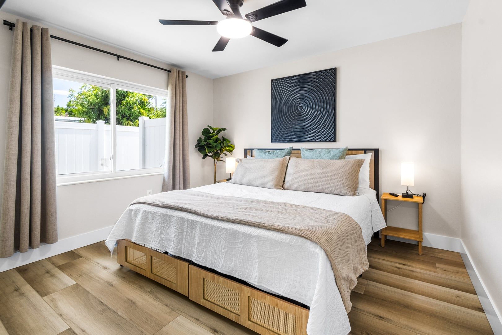 Un dormitorio con cama, ventilador de techo, una obra de arte en la pared, mesita de noche con lámpara y ventana con cortinas.
