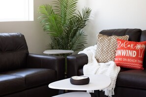 A modern living room with a dark leather sofa, a white coffee table, and a small side table. There is a large green plant in the background and a decorative pillow with a beach theme. - Beachside Bungalow - Unit A | 2BR, 8-Min Walk to Beach, Patio & BBQ Grills 🌴 (Cape Canaveral)