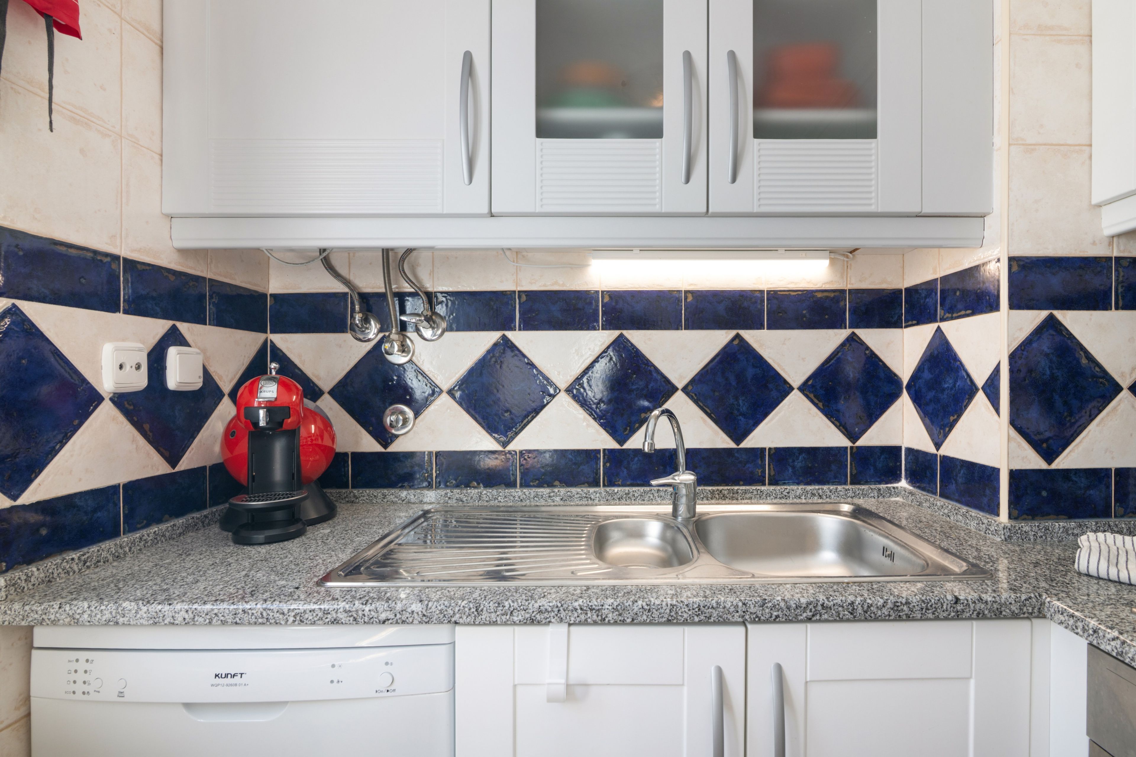A kitchen with white cabinets, a stainless steel sink, a red coffee maker, and blue and white patterned tiles.