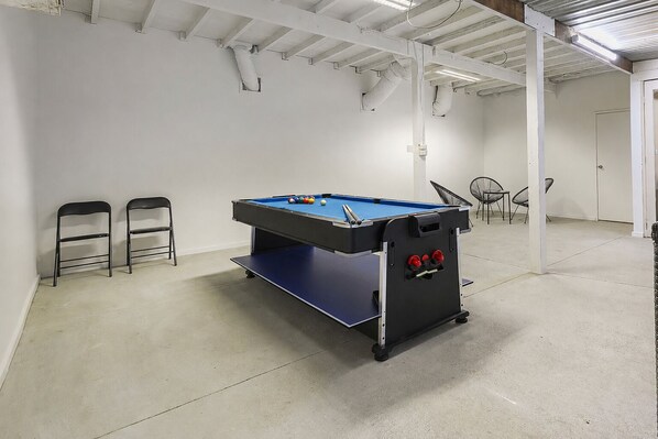 A pool table in a room with white walls and a concrete floor. There are two chairs and a table with chairs in the background.