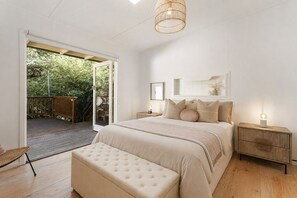 A bedroom with a large bed, a bedside table, a nightstand, a bench, and a door leading to an outdoor area. - Kallista Silvermist House by Ready Set Stay (Kallista)