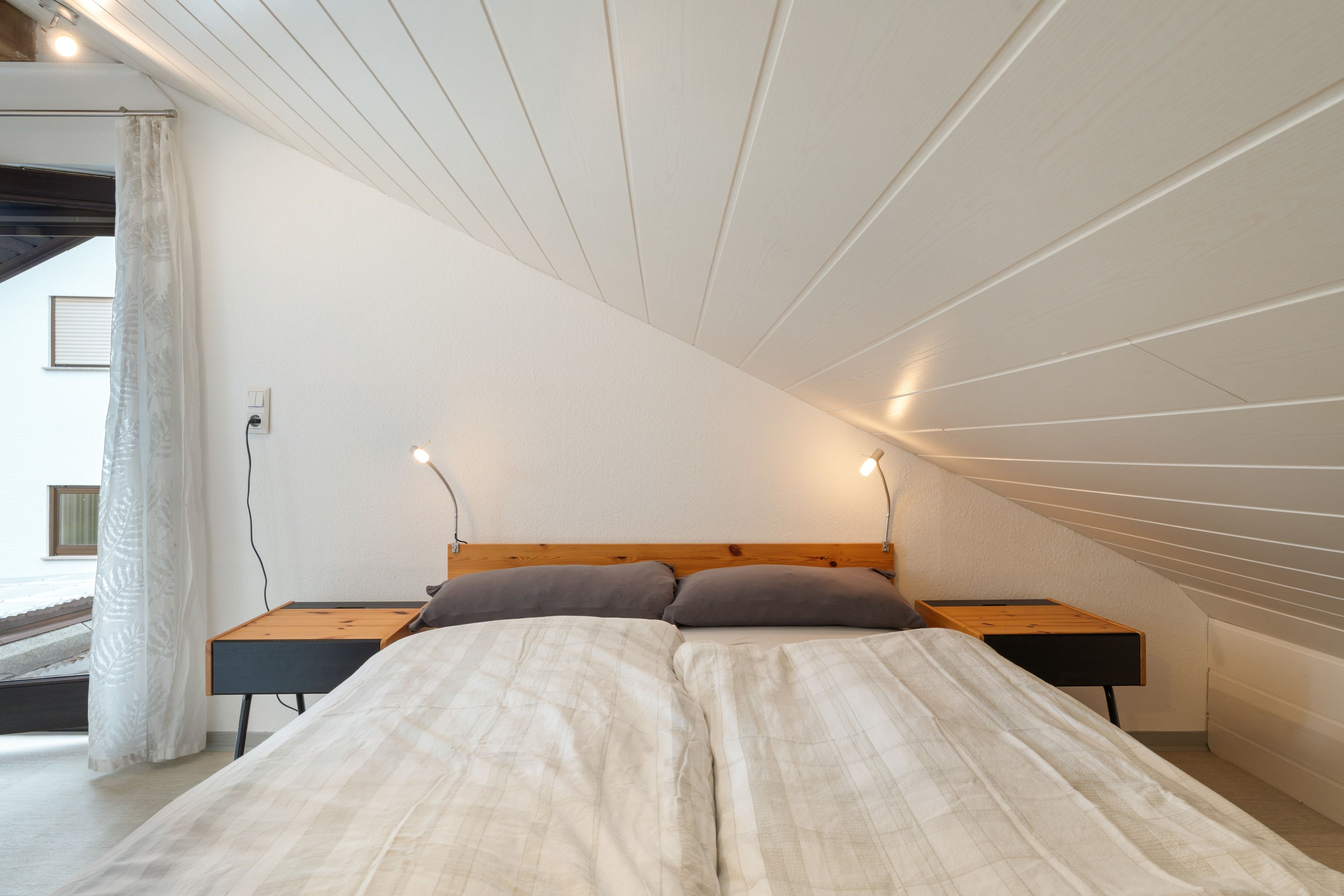 Ein Schlafzimmer mit schrägem Dach, einem Bett mit Holz-Kopfschild, zwei Nachttischen und einem Fenster mit Vorhang.