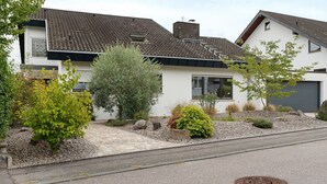 A residential house with a tiled roof, a front yard with various shrubs, and a paved driveway.