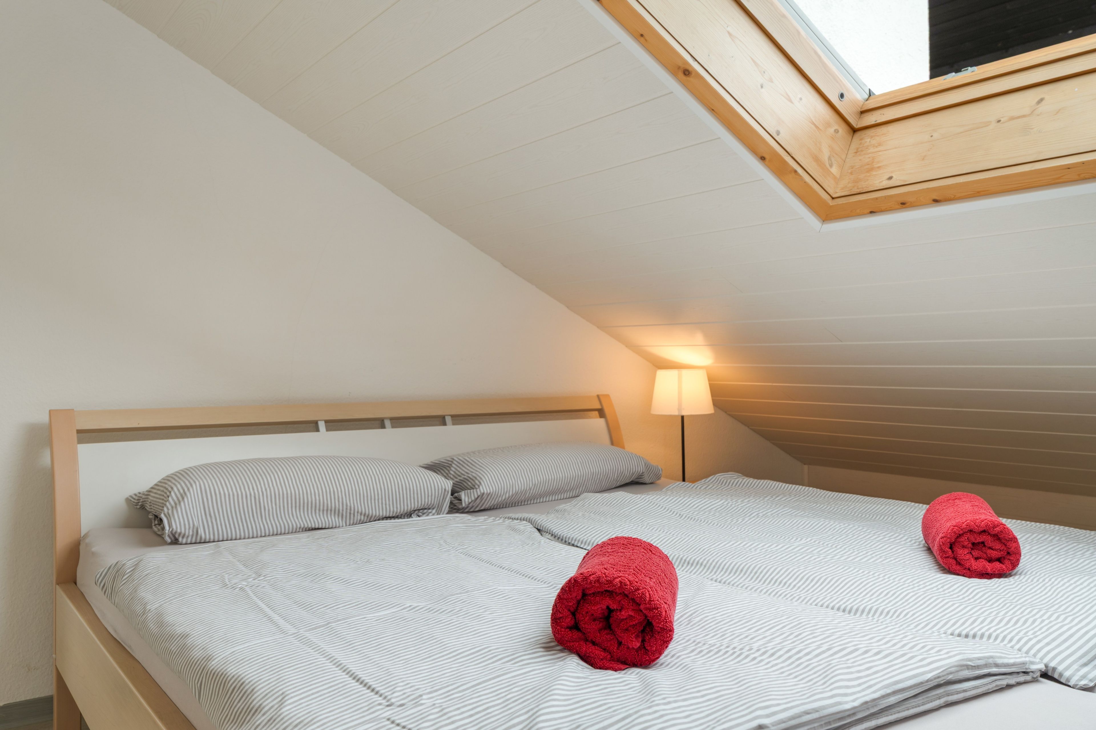 Uma cama de solteiro com cabeceira de madeira, dois travesseiros e dois rolinhos de toalha.