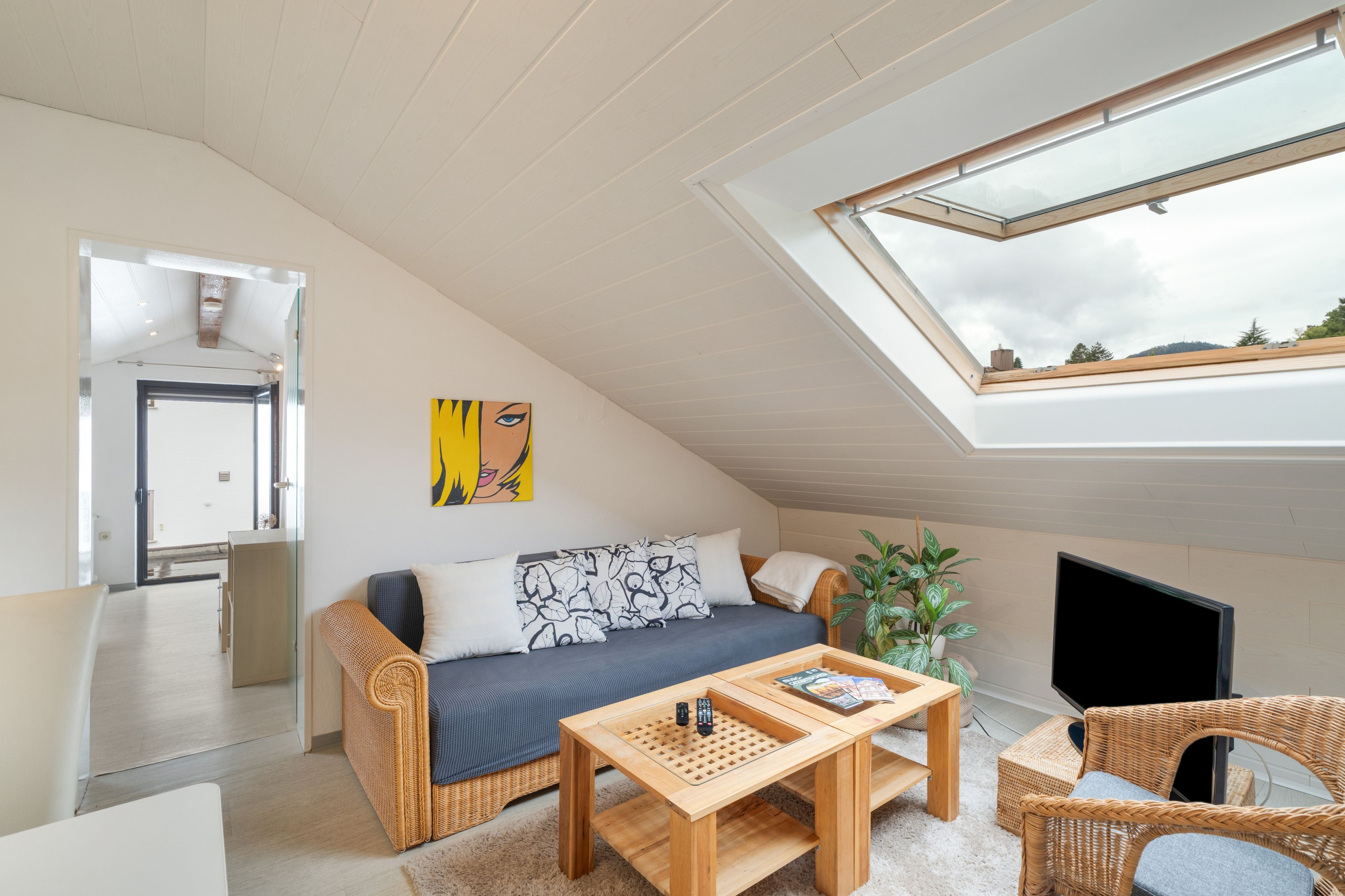 Ein gemütliches Schlafzimmer im Loft-Stil mit schrägem Dach, einer Couch, einem Couchtisch, einem Fernseher und einem großen Fenster.