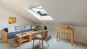 A cozy loft bedroom with a skylight, a sofa, a coffee table, a wicker chair, and a small wooden desk.