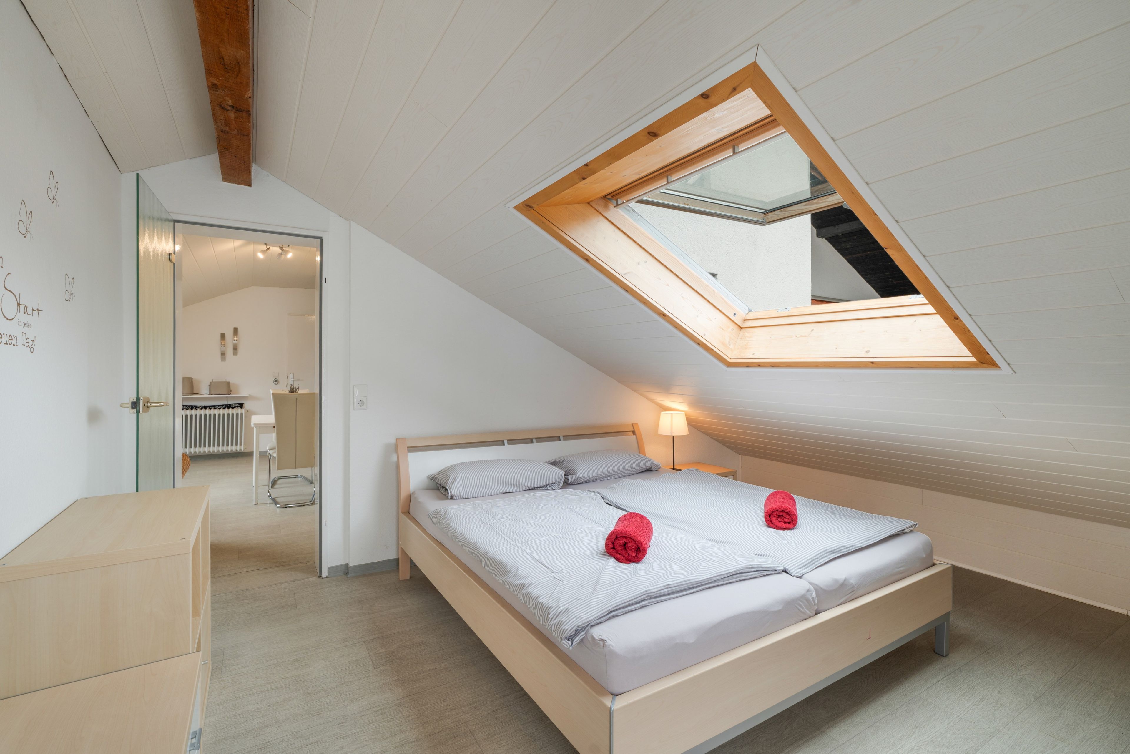 A bedroom with a sloped ceiling, a skylight, a bed with pillows, a bedside lamp, and a small desk.