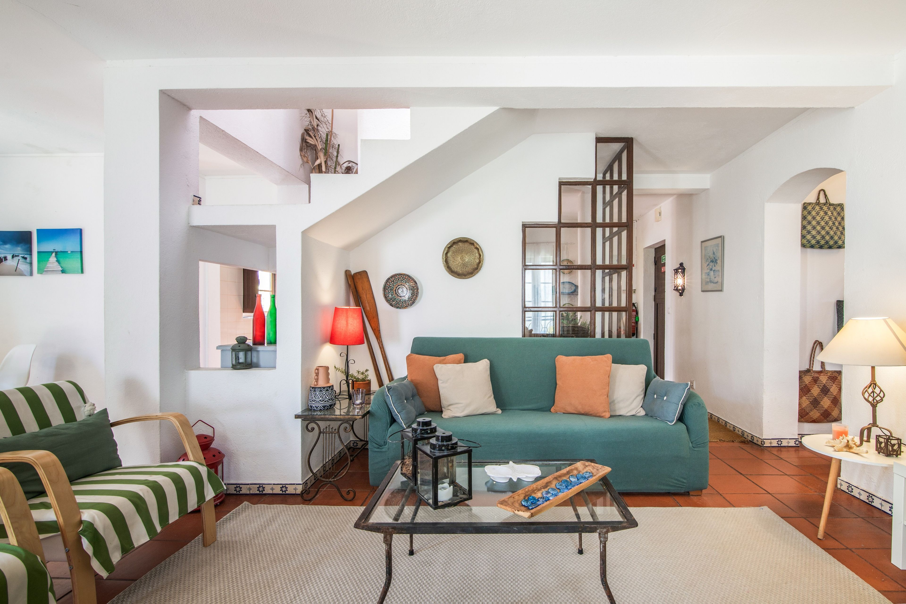 Una sala de estar con un sofá verde, una silla a rayas, una mesa de centro de vidrio y una escalera que lleva a un nivel superior.