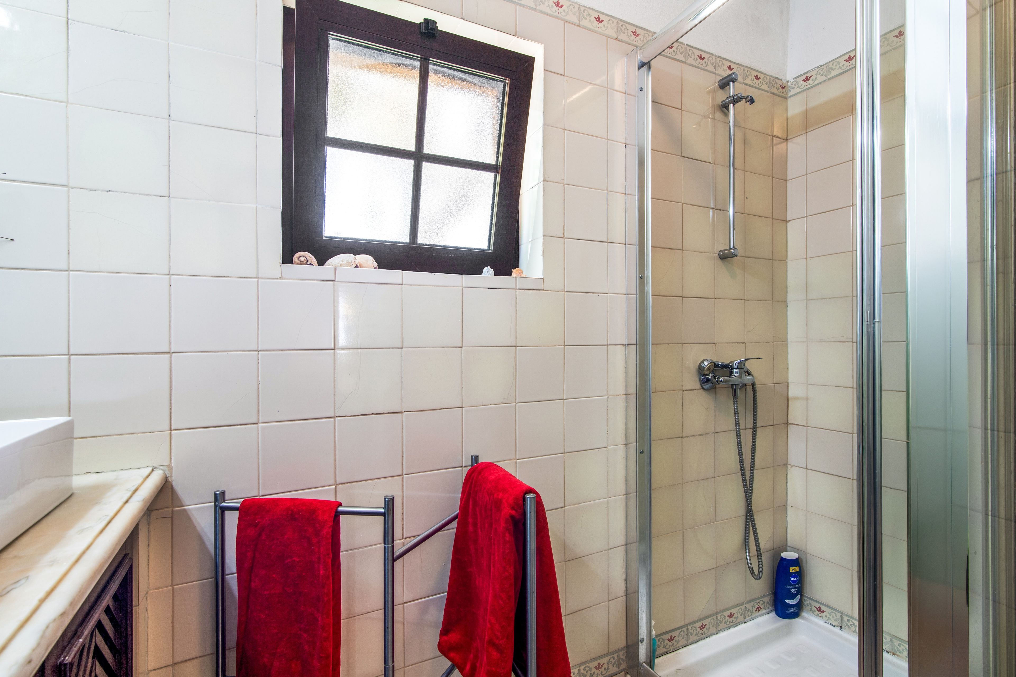 Een badkamer met een douche, een raam en een handdoekrek met twee rode handdoeken.