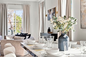 Ein Essbereich mit einem langen Tisch, der für eine Mahlzeit gedeckt ist, einer Vase mit weißen Blumen und Blick ins Freie durch große Fenster.
