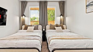 A double room with two beds, a window with curtains, a view of greenery outside, and a painting on the wall. - Luxe 6BR Duplex | Rooftop, Hot Tubs, Games & Views (Dallas)