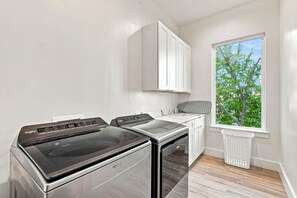 Un lavadero con lavadora y secadora, una ventana con vista a árboles, y un armario blanco sobre el mostrador.