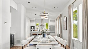 A modern dining room with a long table set for six, a kitchen in the background, and large windows with curtains. - Modern Dallas Retreat | Rooftop, Hot Tub & Views (Dallas)