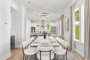 Uma sala de jantar moderna com uma mesa comprida posta para seis pessoas, cozinha ao fundo e grandes janelas com cortinas.