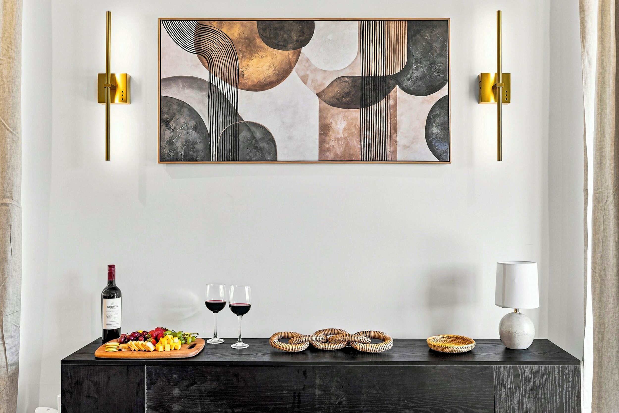 Un intérieur moderne avec une console en bois foncé sur laquelle sont disposés une bouteille de vin, deux verres, une assiette de fruits et un panier tressé. Au-dessus, un grand tableau abstrait est accroché au mur, flanqué de deux appliques murales.