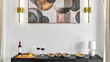 Un intérieur moderne avec une console en bois foncé sur laquelle sont disposés une bouteille de vin, deux verres, une assiette de fruits et un panier tressé. Au-dessus, un grand tableau abstrait est accroché au mur, flanqué de deux appliques murales.