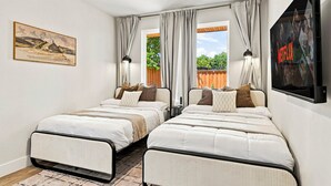 A bedroom with two beds, a ceiling fan, a painting, a window with curtains, and a wall-mounted TV. - Luxe Dallas Home w/ Rooftop, Hot Tub, Yard & Views (Dallas)