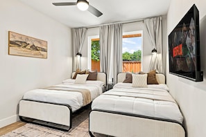 Quarto com duas camas, ventoinha no teto, um quadro, janela com cortinas e televisão presa à parede.
