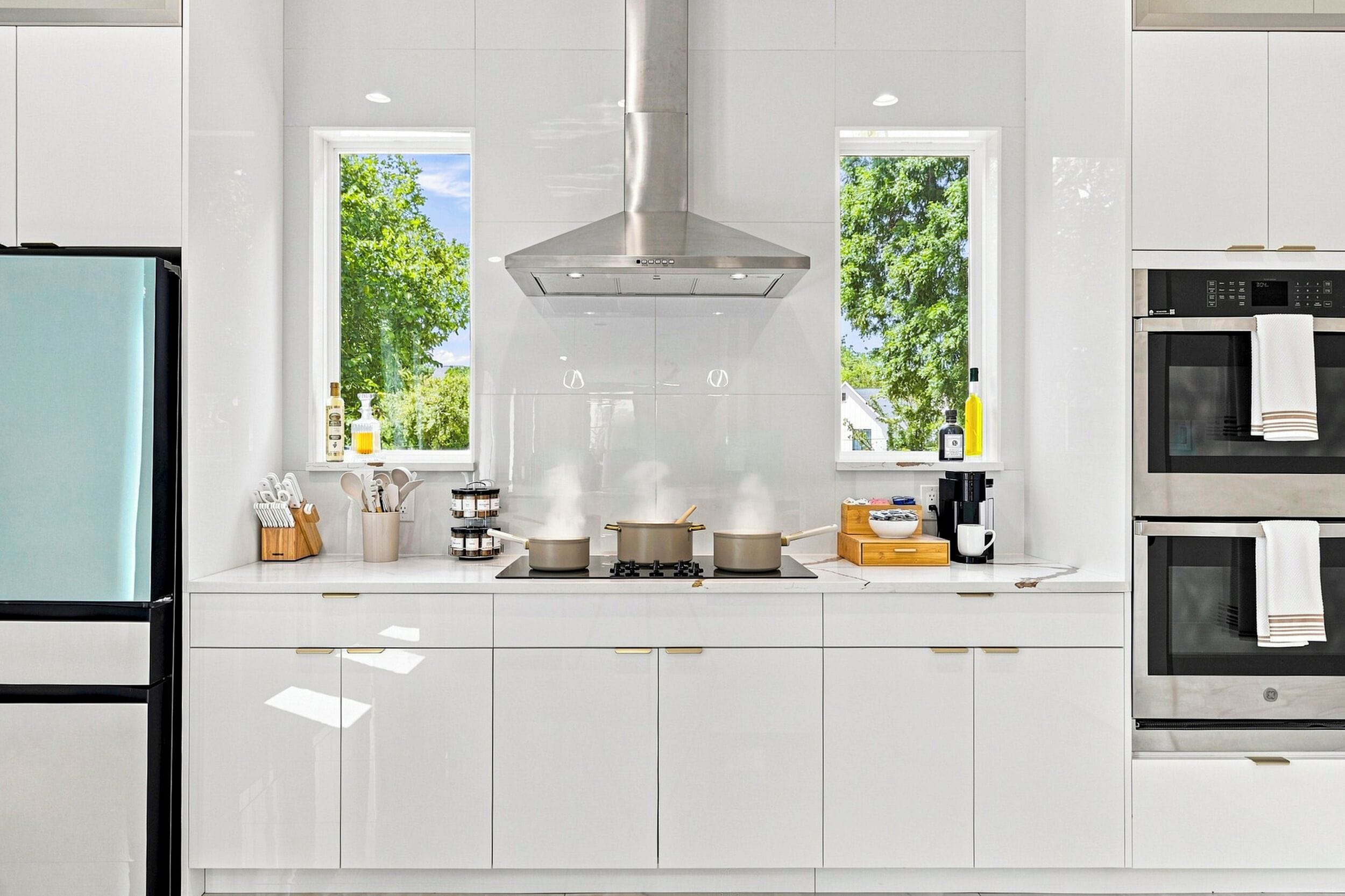 Una cocina moderna con armarios blancos, una campana extractora de acero inoxidable y dos ventanas con vista a árboles.