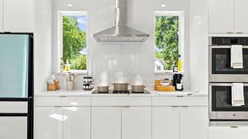 Una cocina moderna con armarios blancos, una campana extractora de acero inoxidable y dos ventanas con vista a árboles.