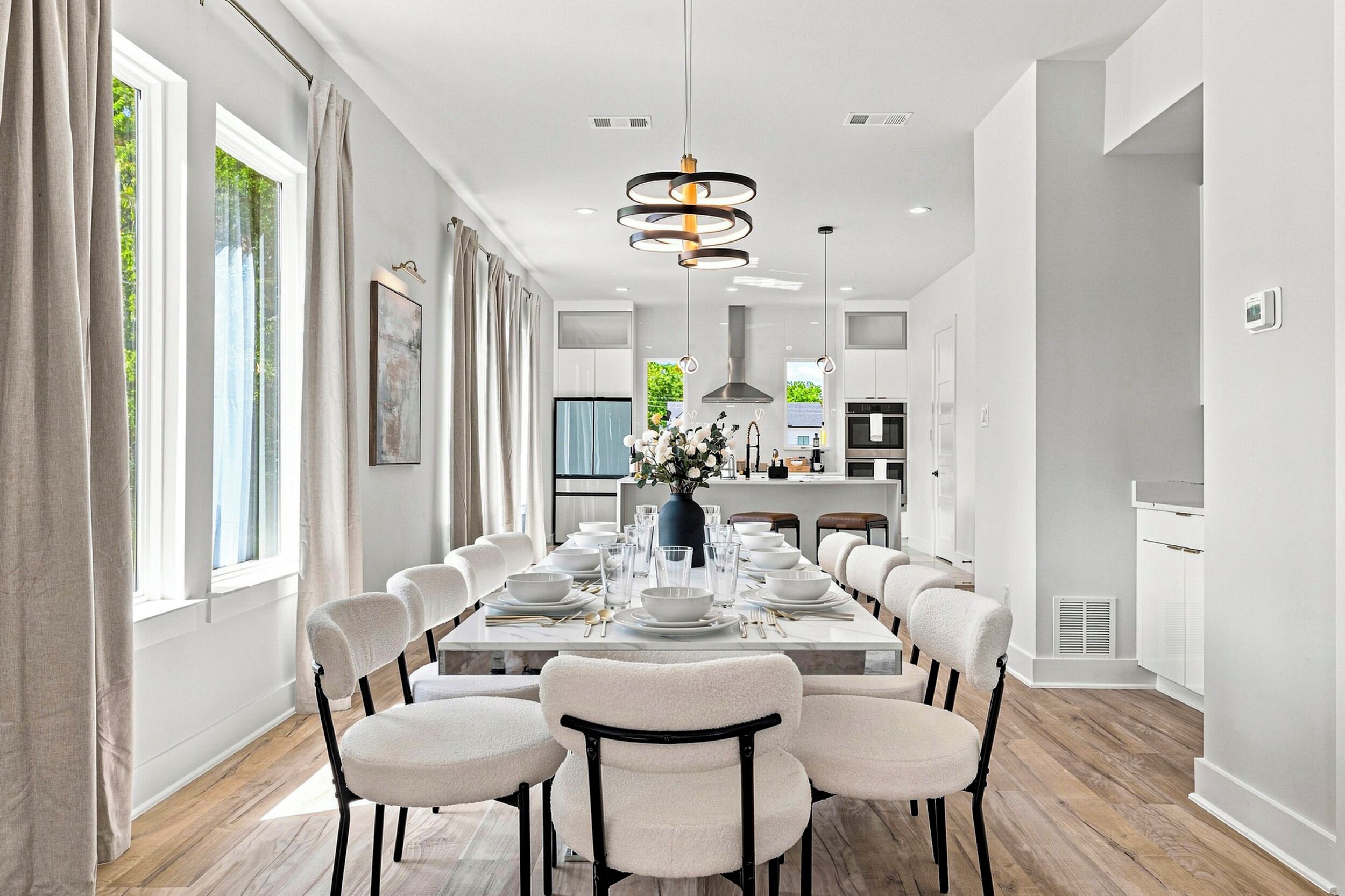 Une salle à manger moderne avec une longue table dressée pour six personnes, entourée de chaises, et une cuisine en arrière-plan.