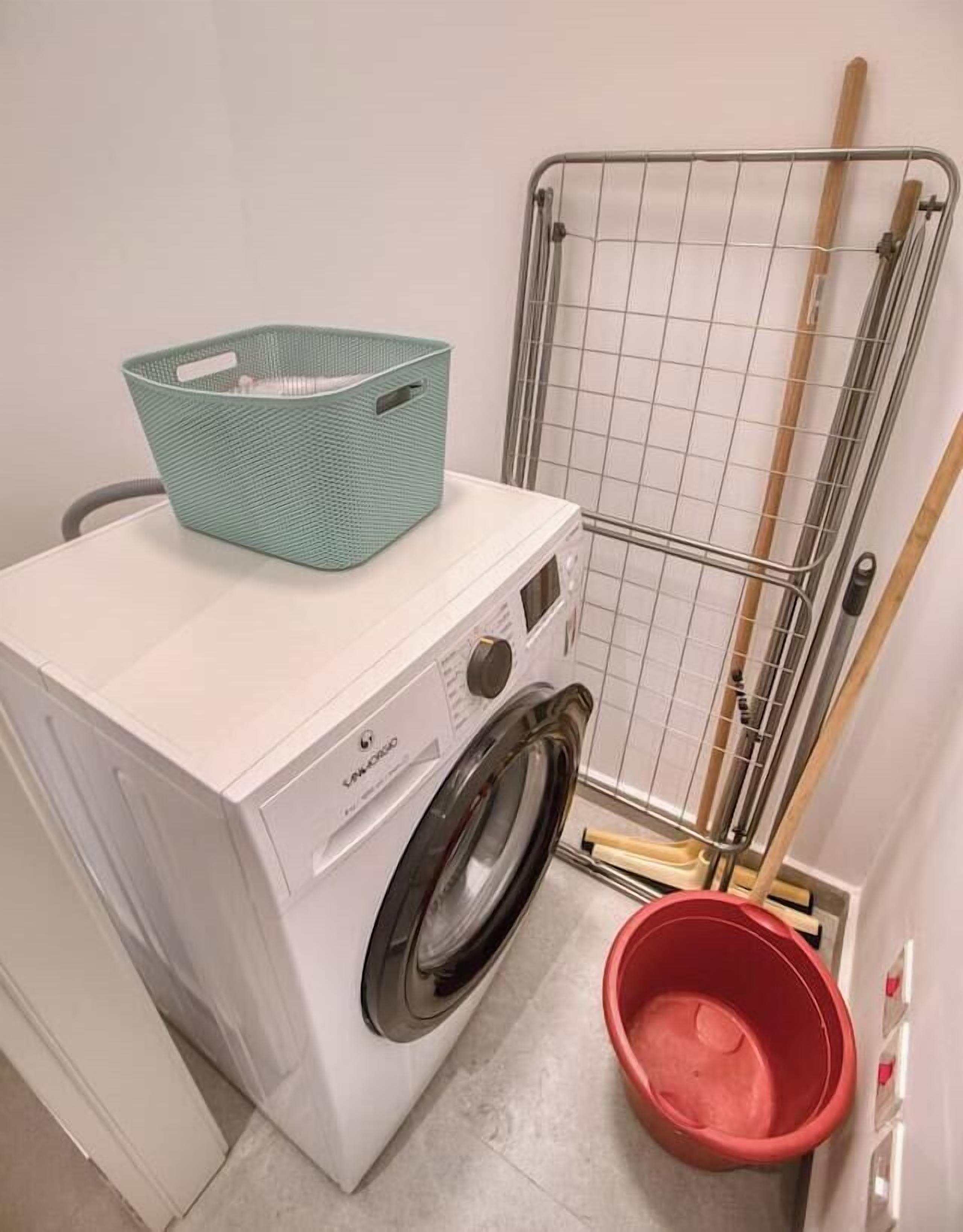 A laundry area with a washing machine, a metal drying rack, and a red bucket.
