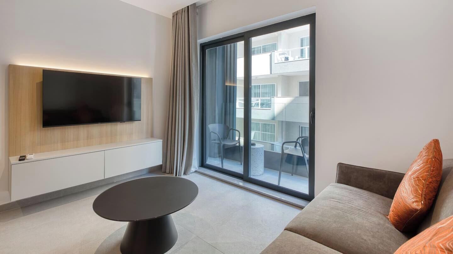 A modern living room with a flat-screen TV mounted on the wall, a round coffee table, a sofa with a cushion, and a large sliding glass door leading to an outdoor balcony.