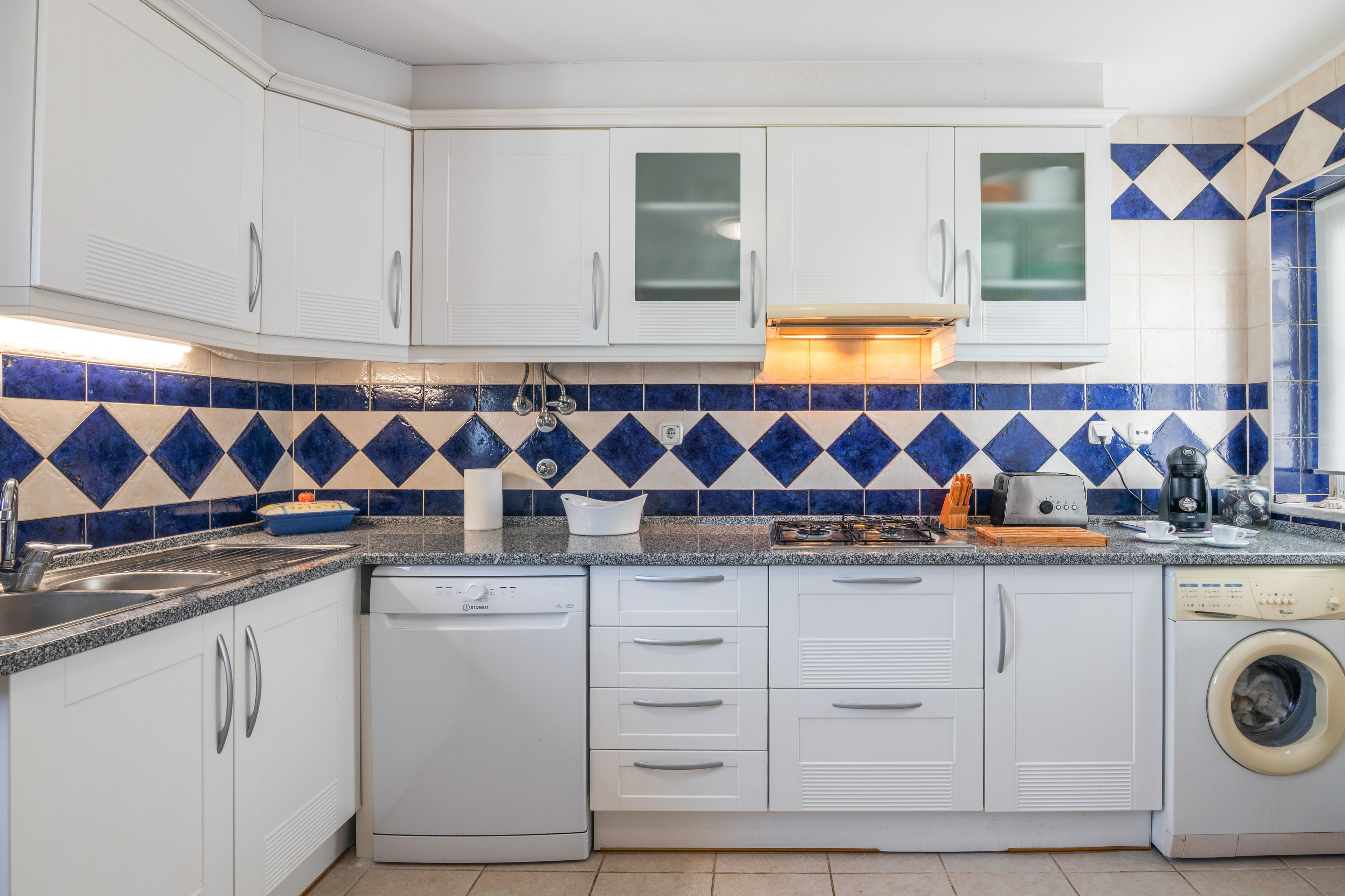 Une cuisine avec des meubles blancs, un lave-vaisselle, un lave-linge et un crédence carrelée aux motifs bleus et beiges.