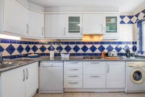 Une cuisine avec des meubles blancs, un lave-vaisselle, un lave-linge et un crédence carrelée aux motifs bleus et beiges.