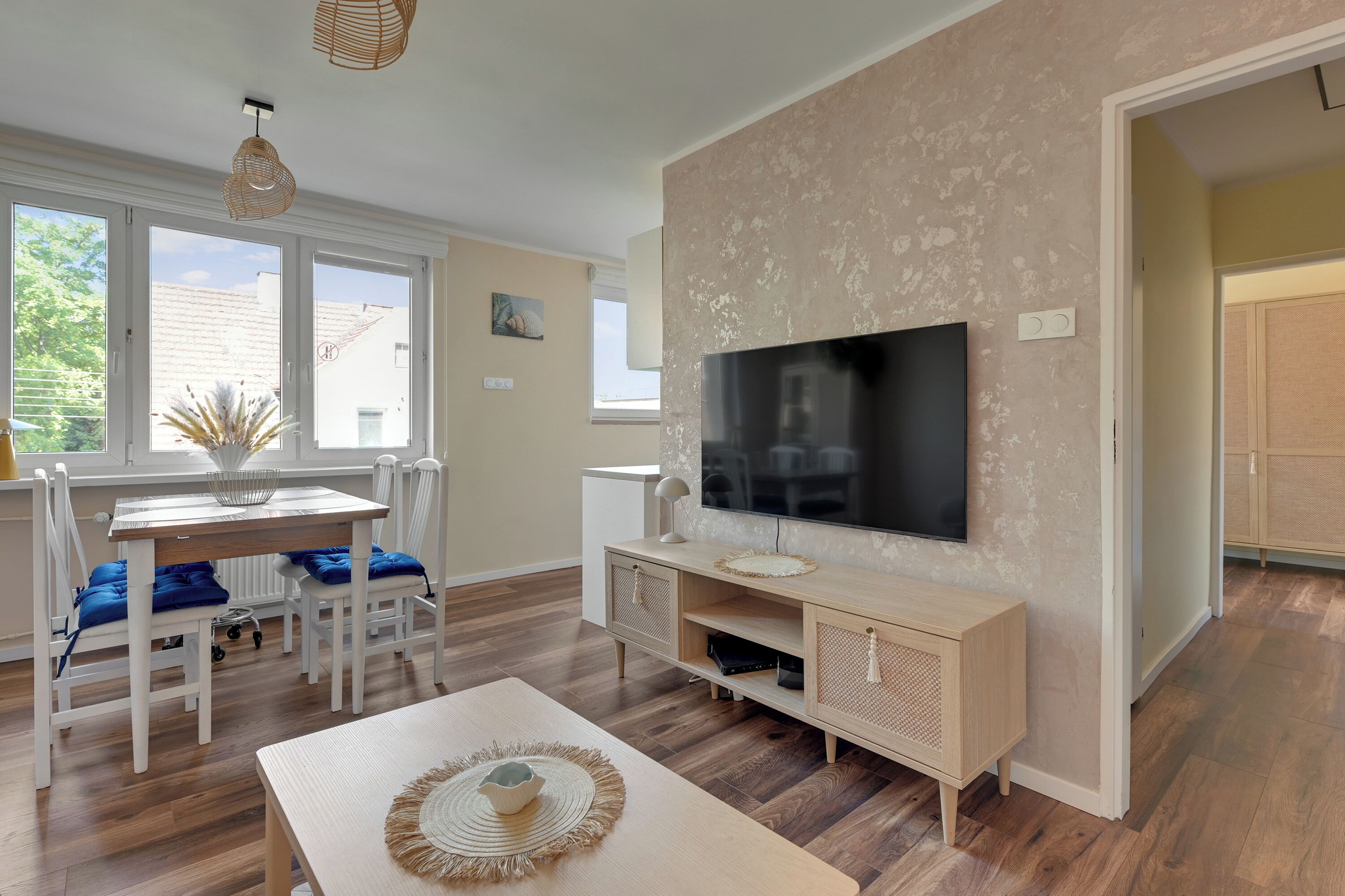A modern living room with a flat-screen TV mounted on the wall, a dining area with a table and chairs, and a large window providing natural light.
