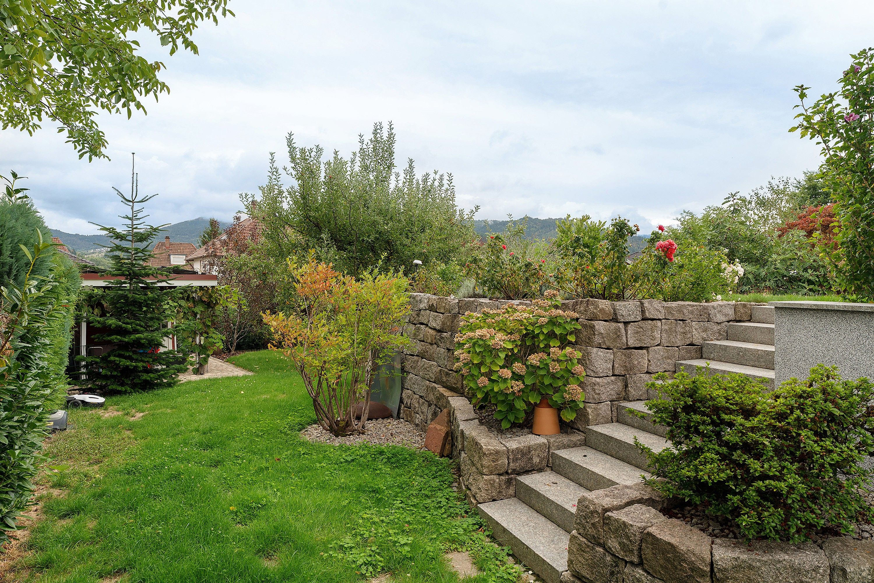Eine Steintreppe führt zu einem Garten mit verschiedenen Pflanzen und Blick auf eine Berglandschaft im Hintergrund.