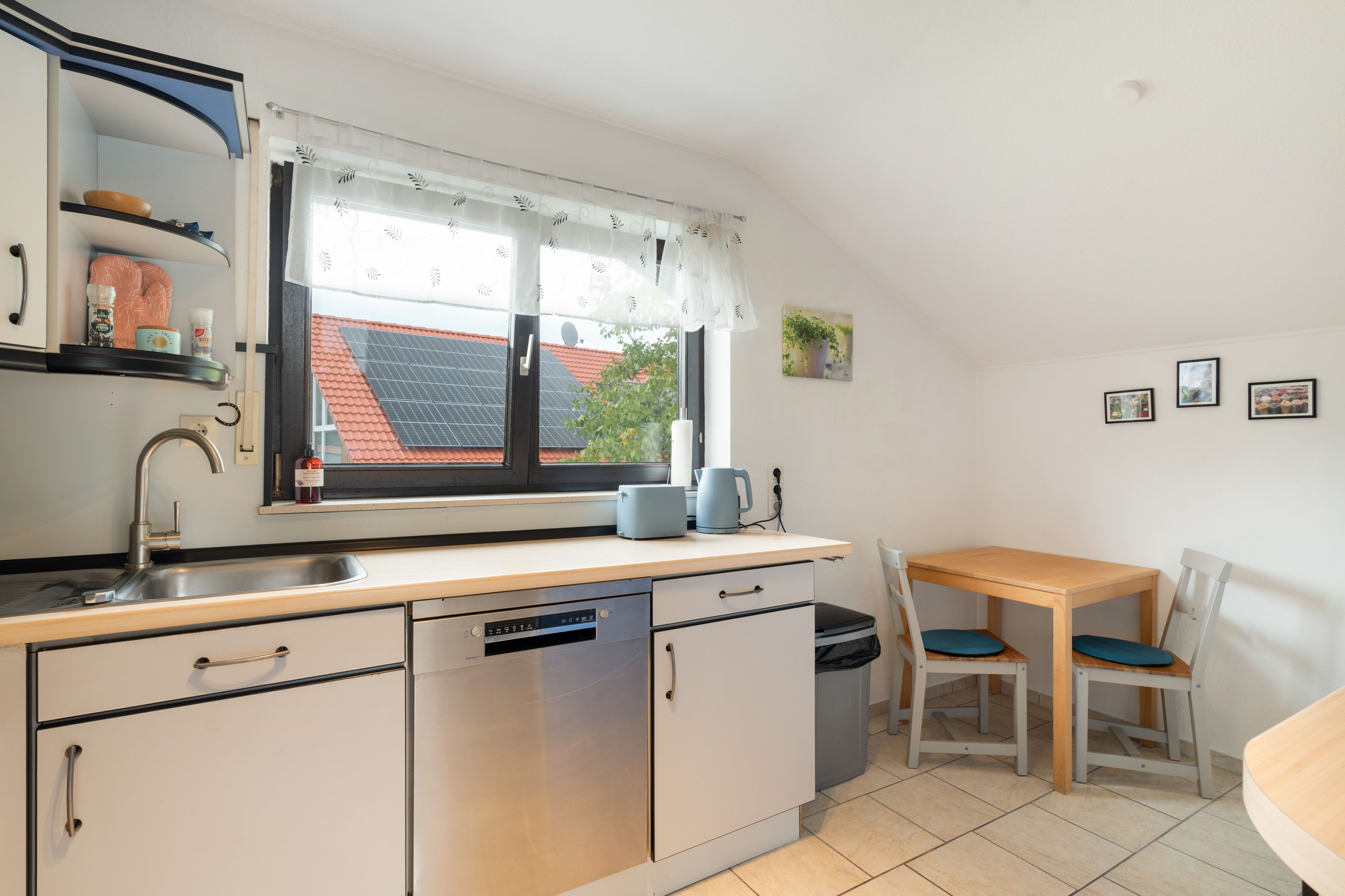 Cocina compacta con lavavajillas, pileta, ventana con vista a una casa, una mesa pequeña con sillas y algunos objetos colgados en la pared.