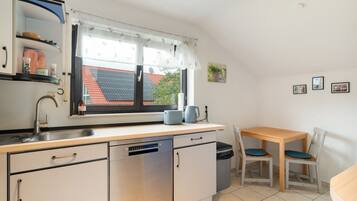 Cocina compacta con lavavajillas, pileta, ventana con vista a una casa, una mesa pequeña con sillas y algunos objetos colgados en la pared.