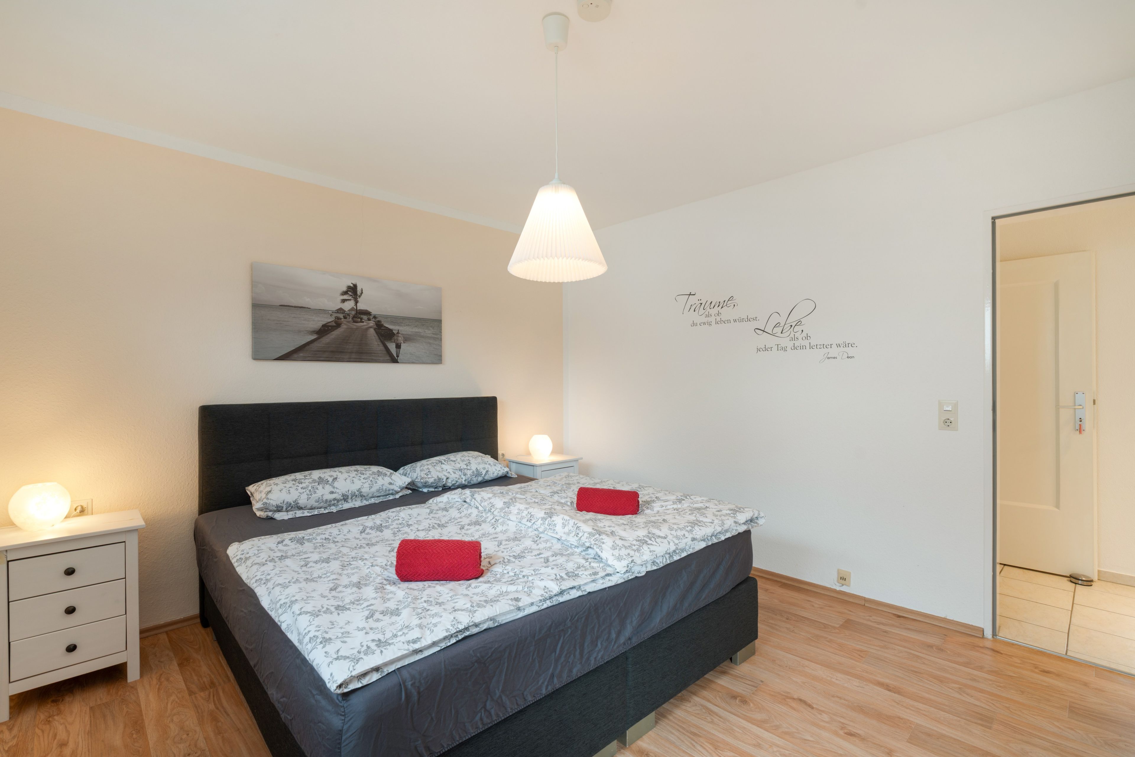 Quarto com cama, mesas de cabeceira, um quadro na parede, luminária pendente e uma porta.