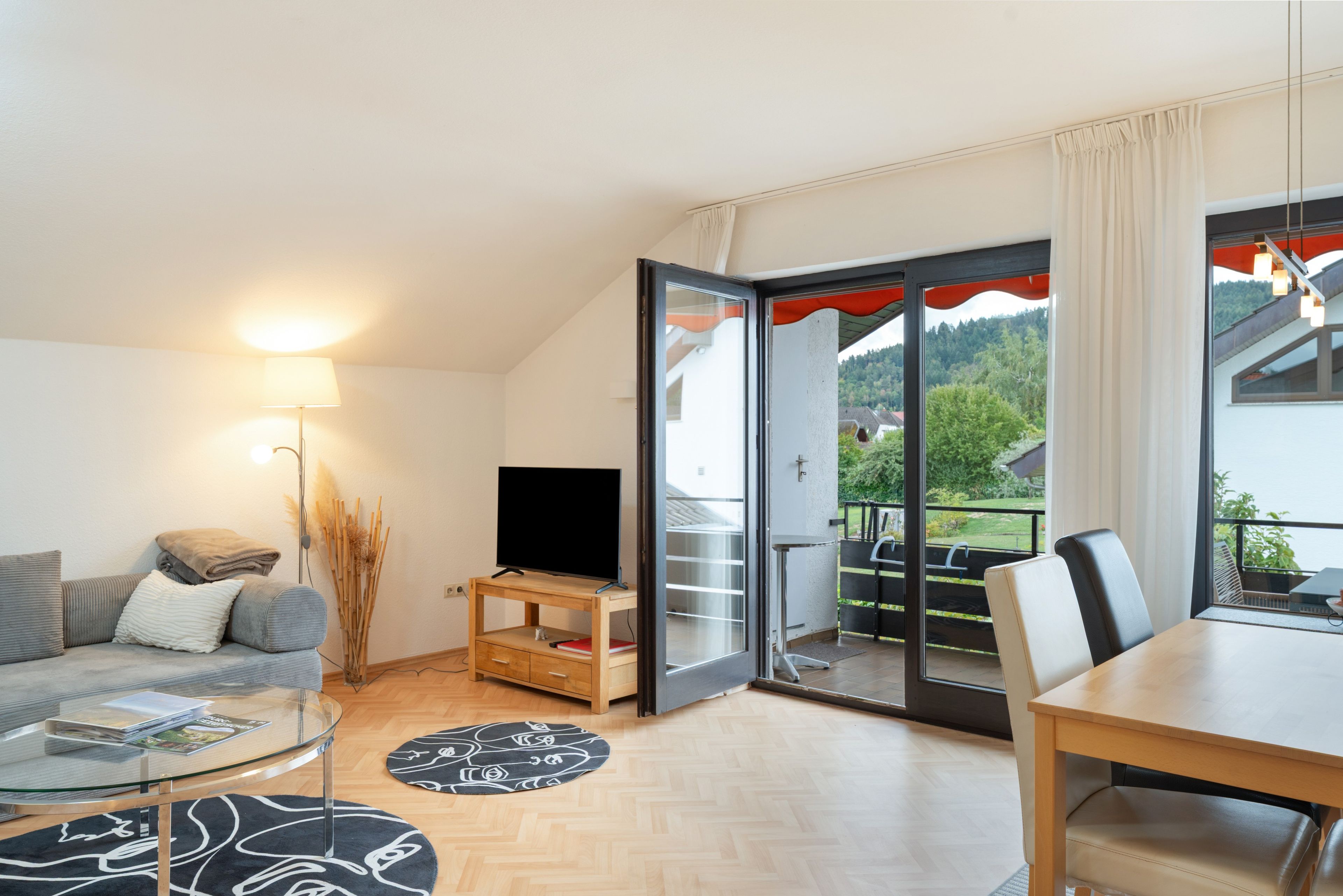 Una sala de estar moderna con un sofá, un televisor sobre un mueble de madera, una mesa de centro de vidrio y una zona de comedor con una mesa y sillas.