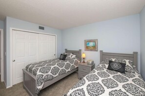 A bedroom with two beds, a nightstand, a lamp, and a door. - Amic 42 2 Bedroom Condo (Bradenton Beach)