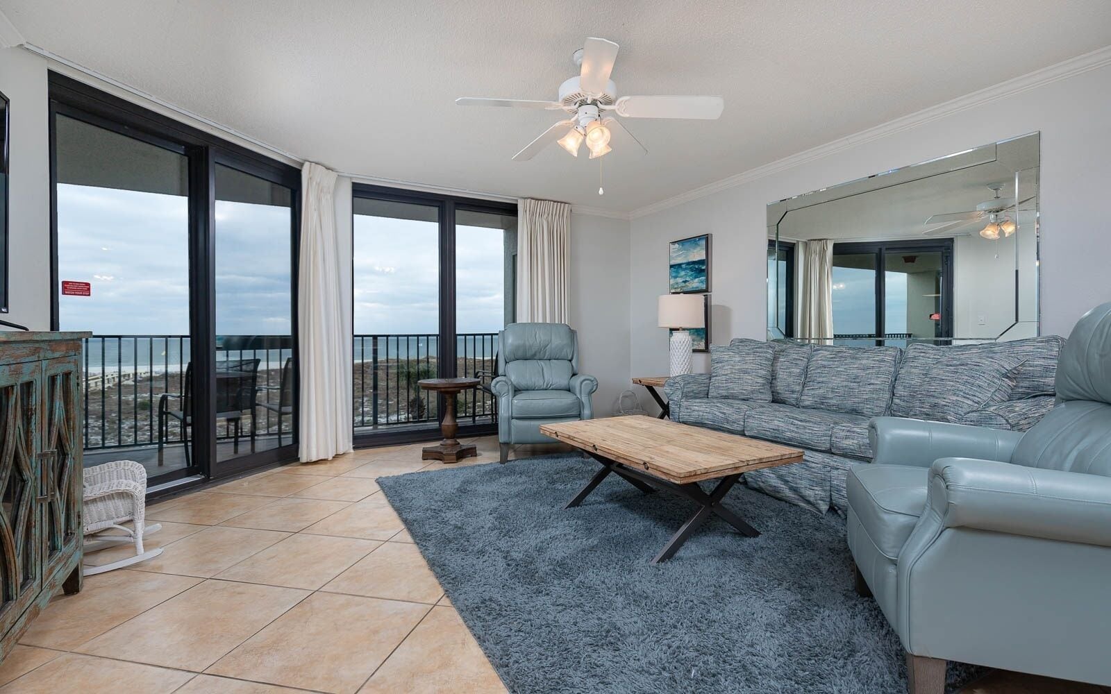 A spacious living room with a large sofa, a coffee table, and a chair. There is a balcony with a view of the beach.