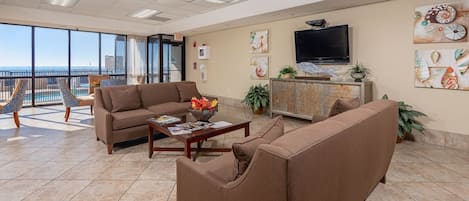 A spacious living room with a large window, a flat-screen TV, and a balcony with a view of the ocean.