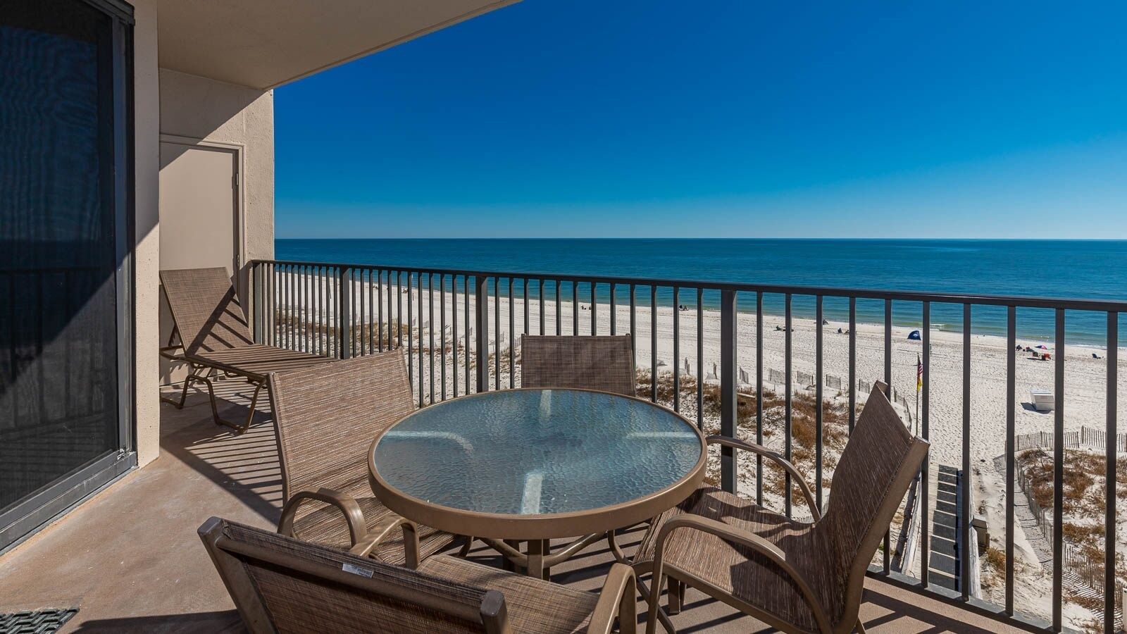 Un balcon avec une table ronde en verre et des chaises en osier, donnant sur une plage avec une vue dégagée sur l’océan.
