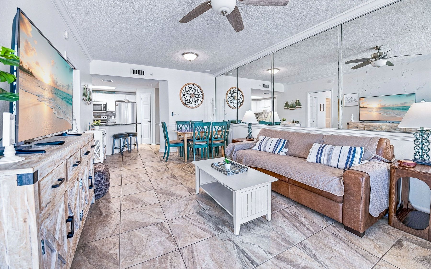 A modern living room with a sofa, coffee table, and a TV. There is a dining area with chairs and a table. The room has a ceiling fan and a large mirror.