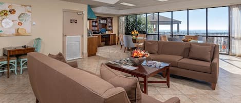 A spacious living area with a large window offering a view of the sea, a comfortable sofa, a coffee table with magazines, and a bookshelf.