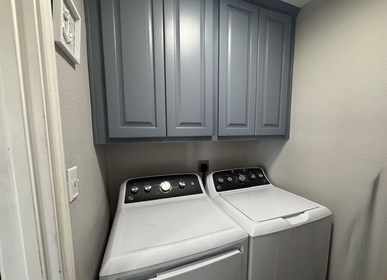 A laundry room with two stacked washers and dryers, grey cabinets above, and a white wall.