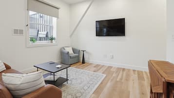 A modern living room with a sofa, a small table, a grey armchair, a TV mounted on the wall, and a wooden floor.