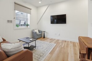 A modern living room with a sofa, a small table, a grey armchair, a TV mounted on the wall, and a wooden floor. - Stylish Downtown Unit With Historic Charm 3 Bedroom Apts (Bloomington)