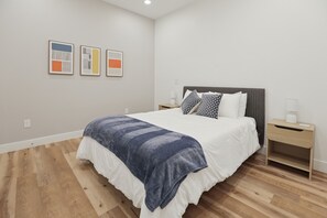 A modern bedroom with a large bed, a bedside table, and a nightstand. There are three framed artworks on the wall. - Stylish Downtown Unit With Historic Charm 3 Bedroom Apts (Bloomington)